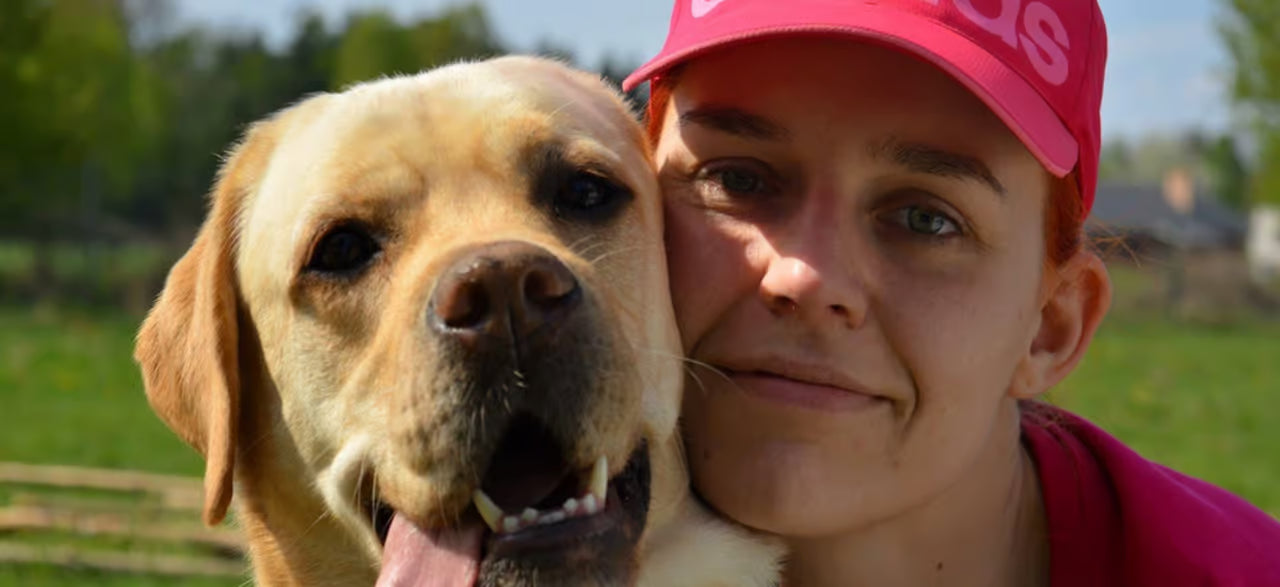 Smiling woman in pink cap hugging a happy Labrador retriever outdoors