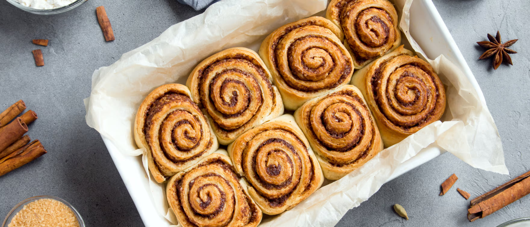 Freshly baked cinnamon rolls in a baking dish, with cinnamon sticks and spices