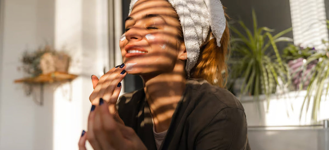 A smiling woman in the morning light applying cream to herself wearing a bathrobe and a turban on her head.