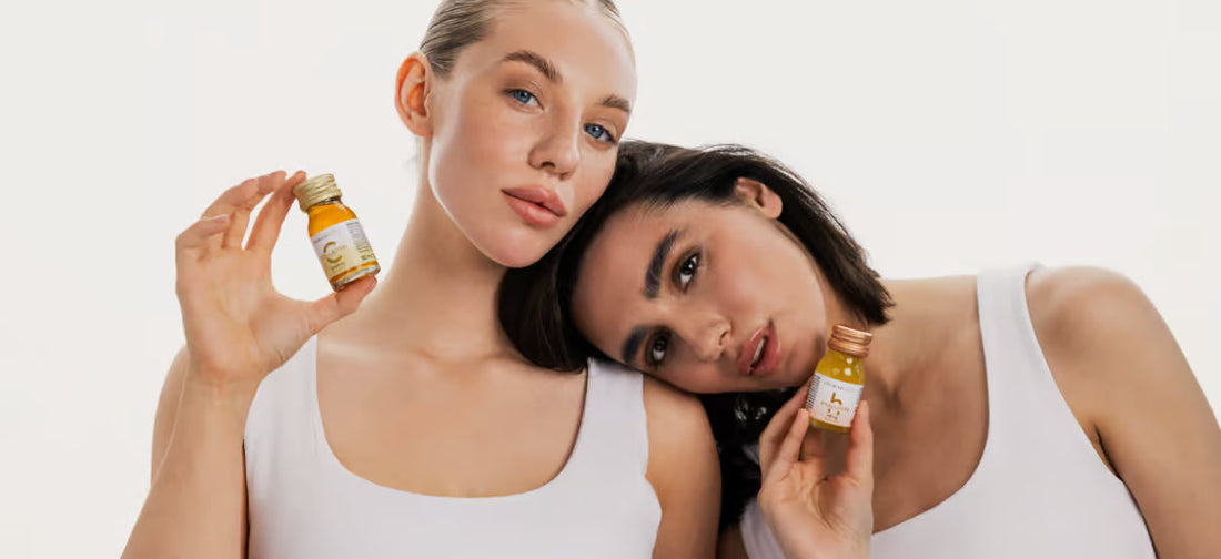 A blonde and a brunette with glowing complexions stand leaning against each other, holding bottles of primabiotic collagen and hyaluronic acid in their hands.