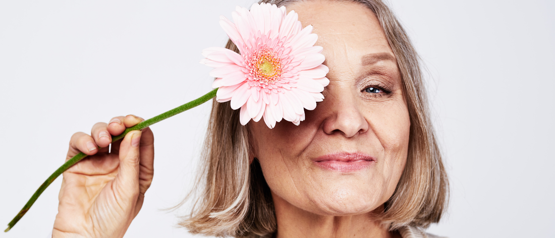 Smiling woman in menopause holding a pink flower over one eye, showing radiant skin and healthy hair after taking liquid collagen Primabiotic