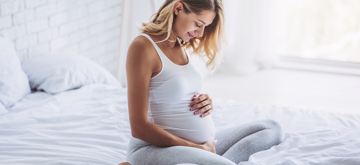 Pregnant woman sitting on bed and gently touching her belly with a smile