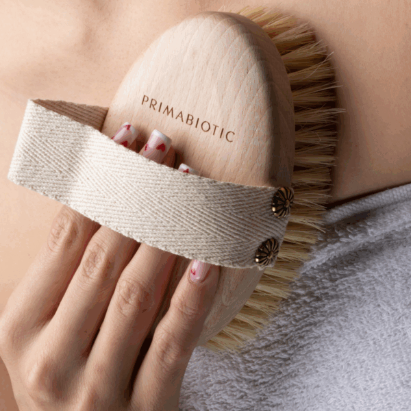 Hand holding Primabiotic wooden body brush with natural bristles and fabric strap, used for dry massage on skin