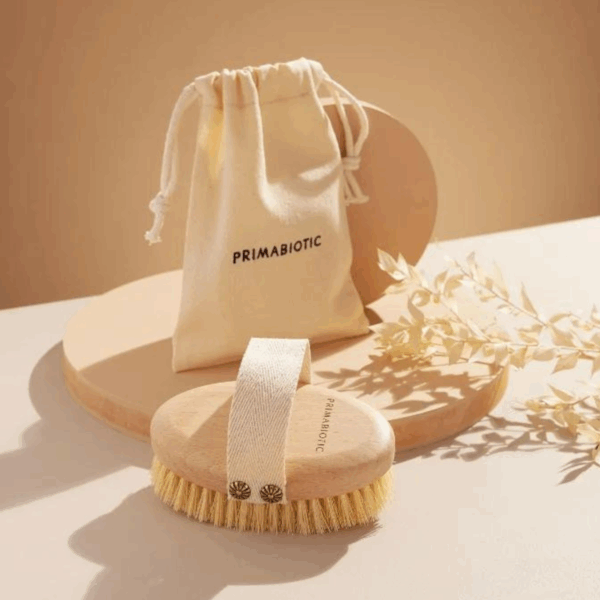 Primabiotic wooden body brush with natural bristles and fabric strap placed beside beige storage pouch with brand logo on light background
