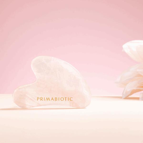 Primabiotic pink gua sha stone with gold logo placed beside light flower on soft pink background, skincare facial massage tool
