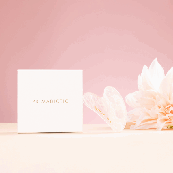 Primabiotic white box and heart-shaped gua sha stone with brand logo placed beside light flower on pink background
